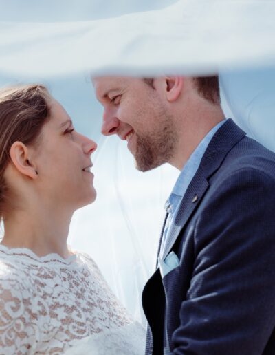 Intimer Moment unter dem Schleier – das Brautpaar lächelt sich verliebt an. Hochzeitsfotografie voller Nähe und Gefühl.