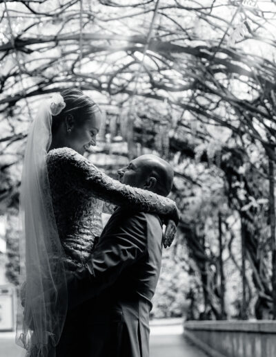 Braut und Bräutigam im Schwarz-Weiß-Porträt unter einem romantischen Blätterdach – er hebt sie mit Leichtigkeit, sie strahlt vor Glück. Klassische Hochzeitsfotografie mit Tiefe.