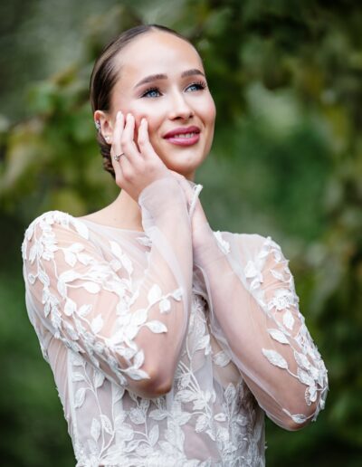 Die Braut im transparenten Spitzenoberteil ihres Kleides steht im Grünen und lächelt verträumt – pure Eleganz und Glück.