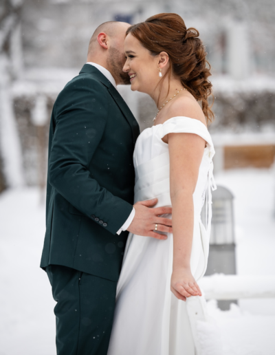 Ein Brautpaar steht in einer Schneelandschaft. Sie lehnt sich an einer Bank und er lehnt sich von vorne an sie ran und flüstert ihr was ins Ohr.