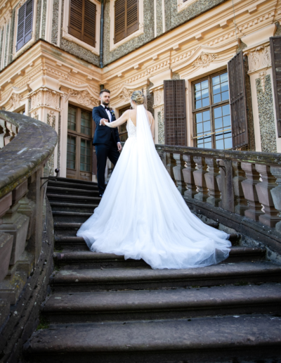 Treppenaufgang zum Schloss, Der Bräutigam steht oben und nimmt die Braut in Empfang, das Kleid der Braut ist ausgebreitet nach unten drapiert.