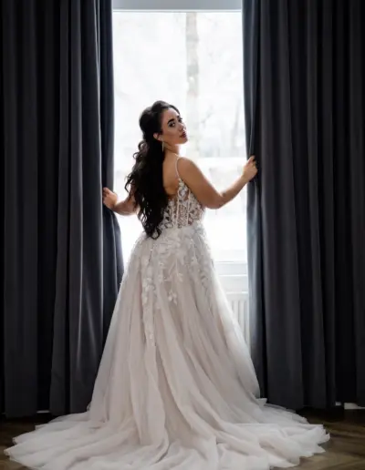 Braut steht im imposanten Brautkleid zwischen zwei dunklen Vorhängen am Fenster. Sie hebt die Vorhänge links und rechts mit ihren Händen fest. Blickt über die Schulter nach hinten.