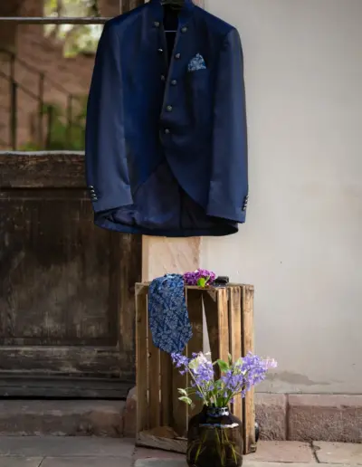 Das Outfit des Mannes hängt auf einem Bügel draussen vor einer Tür. Krawatte liegt unten auf einer Holzbox, Blumen steht in einer Vase daneben.