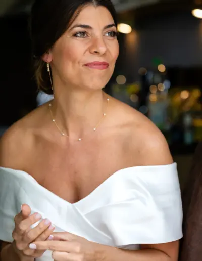 Braut ist bereits angezogen und macht sich gerade noch Parfüm drauf. Schaut glücklich und erwartungsvoll zur Seite. Hinter ihr sind goldene Bokeh Lichter zu sehen.
