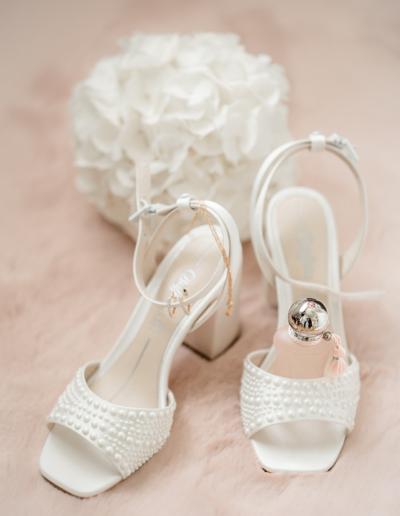 Zwei Brautschuhe stehen auf einem flauschigem rosanem Teppich, in einem Schuh steht eine Parfümflasche mit silbernem Kopf, im anderem Schuhe stecken die Ohrringe und die Kette. Der Brautstrauß ist weiss und im Hintergrund unscharf zu erkennen