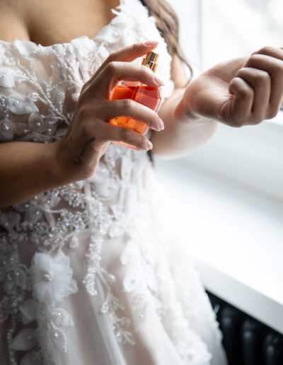 Die Braut steht im Brautkleid am Fenster und trägt sich Parfüm auf das Handgelenk auf. Die Parfümflasche ist orange.