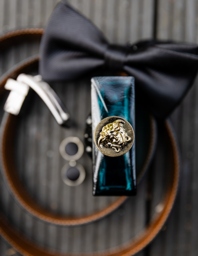 Aufnahme von oben, das Logo von Versace ist von der Parfümflasche zu sehen. Der Gürtel ist unscharf um die Flasche gewickelt, eine Fliege liegt unscharf auf dem Gürtel und die zwei schwarzen Manschettenknöpfe sind unscharf zu sehen.