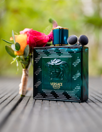 Versace Parfümflasche steht auf einem Holzboden, schwarze Manschettenknöpfe liegen auf ihr. Ein Bunter Blumenanstecker lehnt an der Flasche an