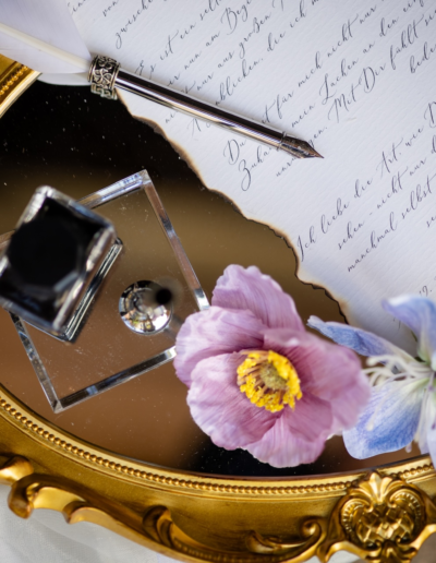 Eine Schreibfeder liegt auf einem beschriebenem Blatt Papier. Tintenfass steht Nebendran und zwei Blumen. liegen seitlich. Alles auf einem goldenem Tablett angerichtet