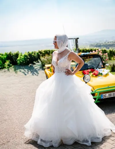 Braut steht mit Sonnenbrille und Kopftuch vor dem Hochzeitsauto. Hände in die Hüften gestemmt und schaut zur Seite.