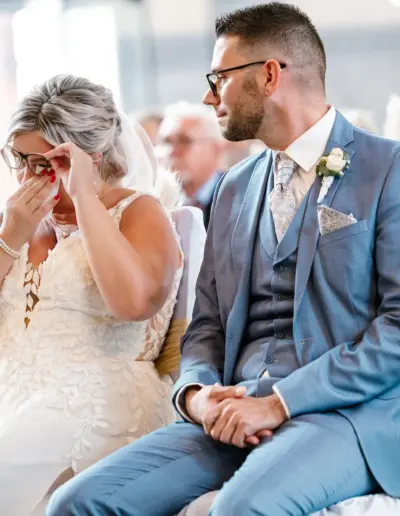 Brautpaar in der Kirche sitzend, Bräutigam schaut verlegen zur Braut, beide Hände im Schoss. Braut wischt sich unter ihrer Brille mit einem Taschentuch eine Träne weg.