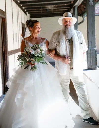 Eine Braut in einem opulentem Kleid hat sich beim Vater untergehackt und blickt lächelnd zu ihm. Der Brautvater blickt stolz Richtung Kamera.