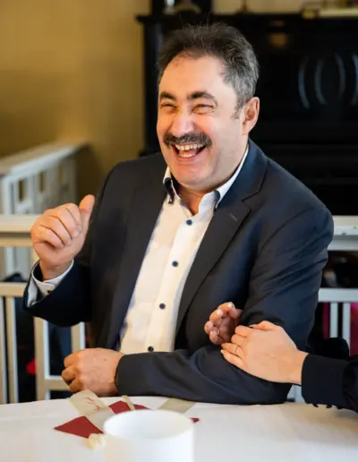 Ein Mann lacht herzlich über etwas und zeigt dabei mit dem Daumen nach hinten. Zwei Frauenhände umfassen seinen Arm.