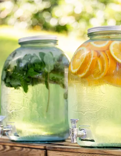 Zwei große Getränkebehälter aus Glas mit vielen Früchten drin stehen in einer grünen Landschaft.