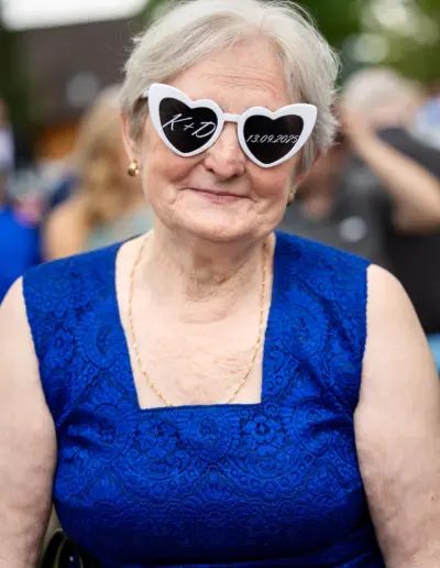 Ein ältere Frau sitzt da und schaut zur Kamera, sie trägt eine Herzbrille mit dem Datum und Initialen des Brautpaares.