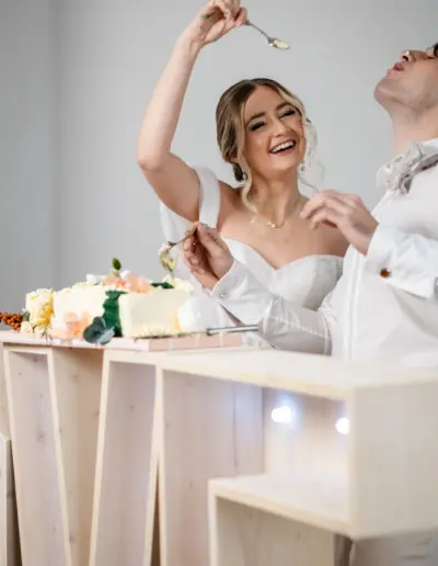 Braut füttert mit einem kleinem Löffel den Bräutigam mit einem stück Kuchen. Sie lacht dabei und der Bräutigam hat den Kopf nach hinten geworfen. Auf seiner Nase ist ein Klecks Kuchen.